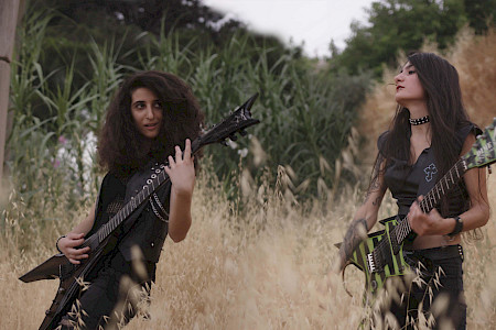 Zwei Personen mit langen Haaren spielen E-Gitarre in einem hohen Grasfeld, umgeben von Büschen und Bäumen.