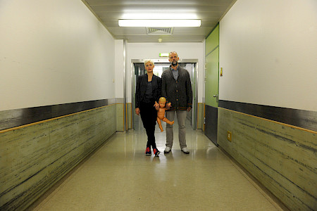 Zwei Personen gehen in einem Krankenhausflur, eine hält eine nackte Babypuppe in der Hand.