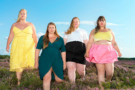 Vier Frauen in Sommerkleidern und Rock stehen auf einer Wiese mit lila blühenden Pflanzen unter blauem Himmel.