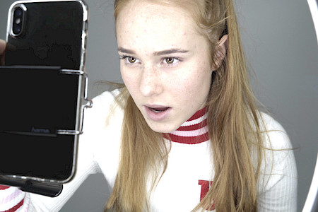 Junge Frau mit langen blonden Haaren trägt weißen Pullover mit roten Streifen und hält ein Smartphone vor sich.