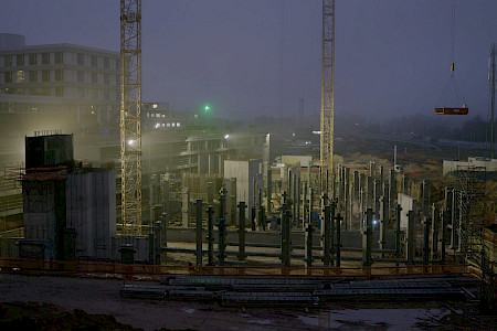 Baustelle mit mehreren Betonpfeilern und zwei hohen Kränen bei Dämmerung oder Nacht, Gebäude im Hintergrund.
