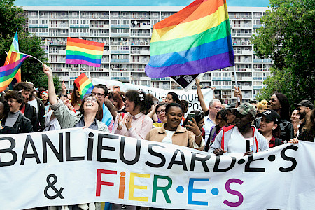 Gruppe von Menschen hält ein Banner mit der Aufschrift 'BANLIEUSARD·E·S & FIER·E·S' und schwenkt Regenbogenfahnen vor einem Wohnblock.