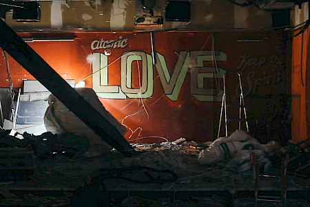 Rotes Wandbild mit dem Schriftzug 'atomic LOVE' in großen weißen Buchstaben, umgeben von Trümmern und einem schräg liegenden Balken.