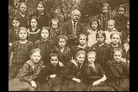 Gruppenfoto mit mehreren Kindern und einem Erwachsenen in historischer Kleidung, sitzend und stehend im Freien vor Bäumen.