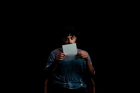 Person sitzt auf einem Stuhl und hält ein weißes Blatt Papier vor sich, dunkler Hintergrund.