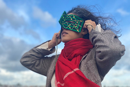 Person mit lockigem Haar, grauem Mantel und rotem Schal, die Hände an den Kopf hebt, vor bewölktem Himmel.