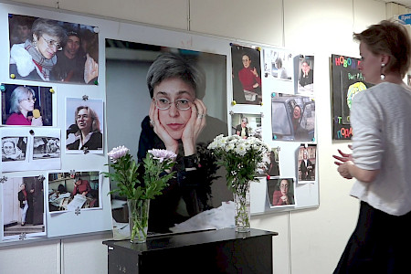 Frau mit kurzem Haar und weißem Pullover steht vor einer Wand mit mehreren Fotos und Postern, auf einem schwarzen Klavier stehen zwei Vasen mit Blumen.