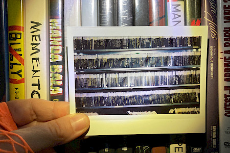 Hand hält ein Foto vor ein Regal mit mehreren DVDs und Blu-rays, darunter Titel wie 'Memento' und 'Armee der Baby Jane'.