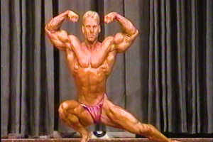 Muscular man with blond hair wearing red briefs poses on stage in front of gray curtain, flexing biceps and thigh muscles.